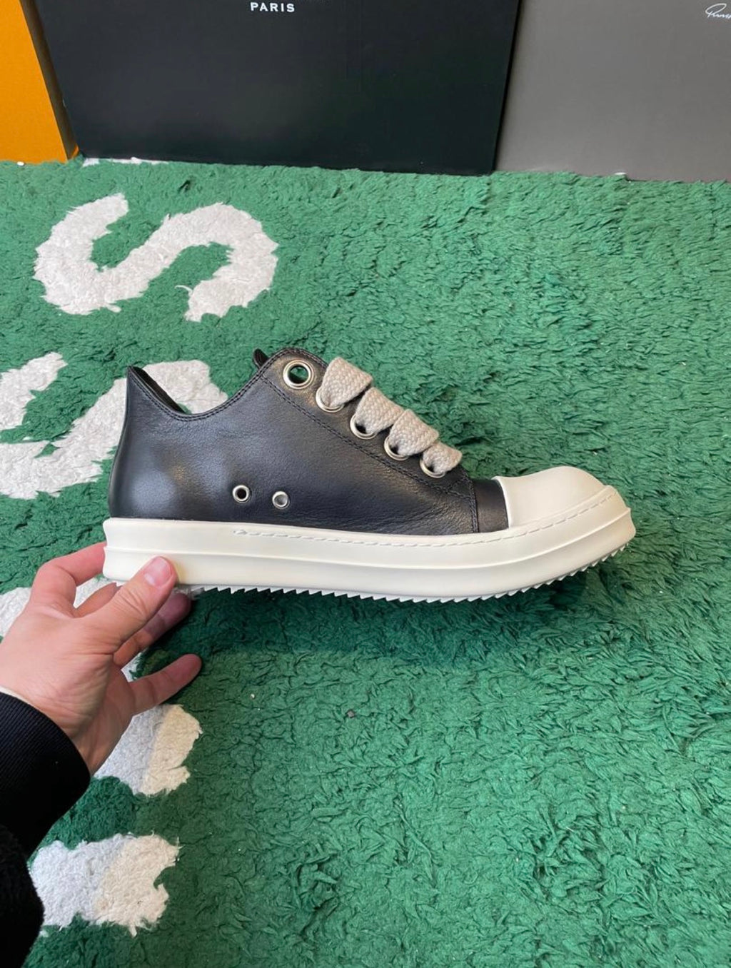 A low-top sneaker with a dark gray leather upper, beige laces, and a thick off-white rubber sole, held by a hand over a green textured rug.